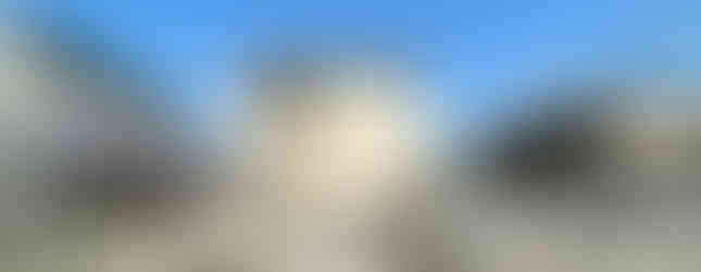 A photo of a two-story house with a white facade and a brown roof. The house features a balcony on the second floor and a carport in the front yard. There is a small garden with a fence around the property. The house is surrounded by other houses and trees, and there are utility poles and wires visible in the background. The sky is blue with some clouds.