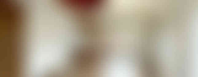 The photograph shows a bright and spacious living room with a wooden floor and white walls. A red circular ceiling decoration adds a touch of color. Large windows offer views of the outside, and a small breakfast bar separates the living area from the kitchen, which is visible in the background.