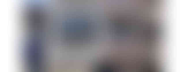 The image shows a three-story house with a gray facade and a brown brick accent wall. The house has a garage on the ground floor, a balcony on the second floor, and a bay window on the third floor. There is a black door and a mailbox in front of the house. The house is located in a residential area with other houses visible in the background.