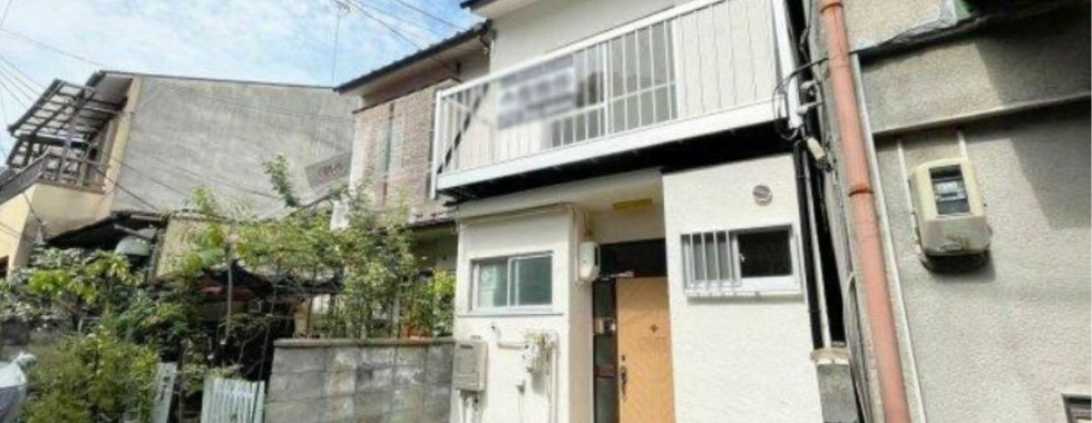 A photo of a two-story house with a white facade and a red tiled roof. The house features a balcony on the second floor and a carport in the front yard. There is a small garden with a fence around the property. The house is surrounded by other houses and trees, and there are utility poles and wires visible in the background. The sky is blue with some clouds.