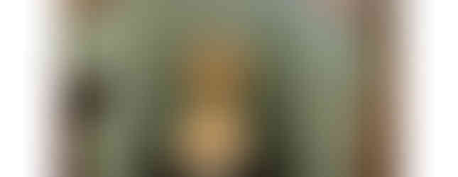 The photograph depicts a small, old-fashioned toilet room with green tiles on the walls. The toilet itself is a vintage beige color with a matching lid. A small sink is attached to the wall above the toilet, and a toilet paper holder is mounted on the adjacent wall. The floor is covered in small, black and white tiles.