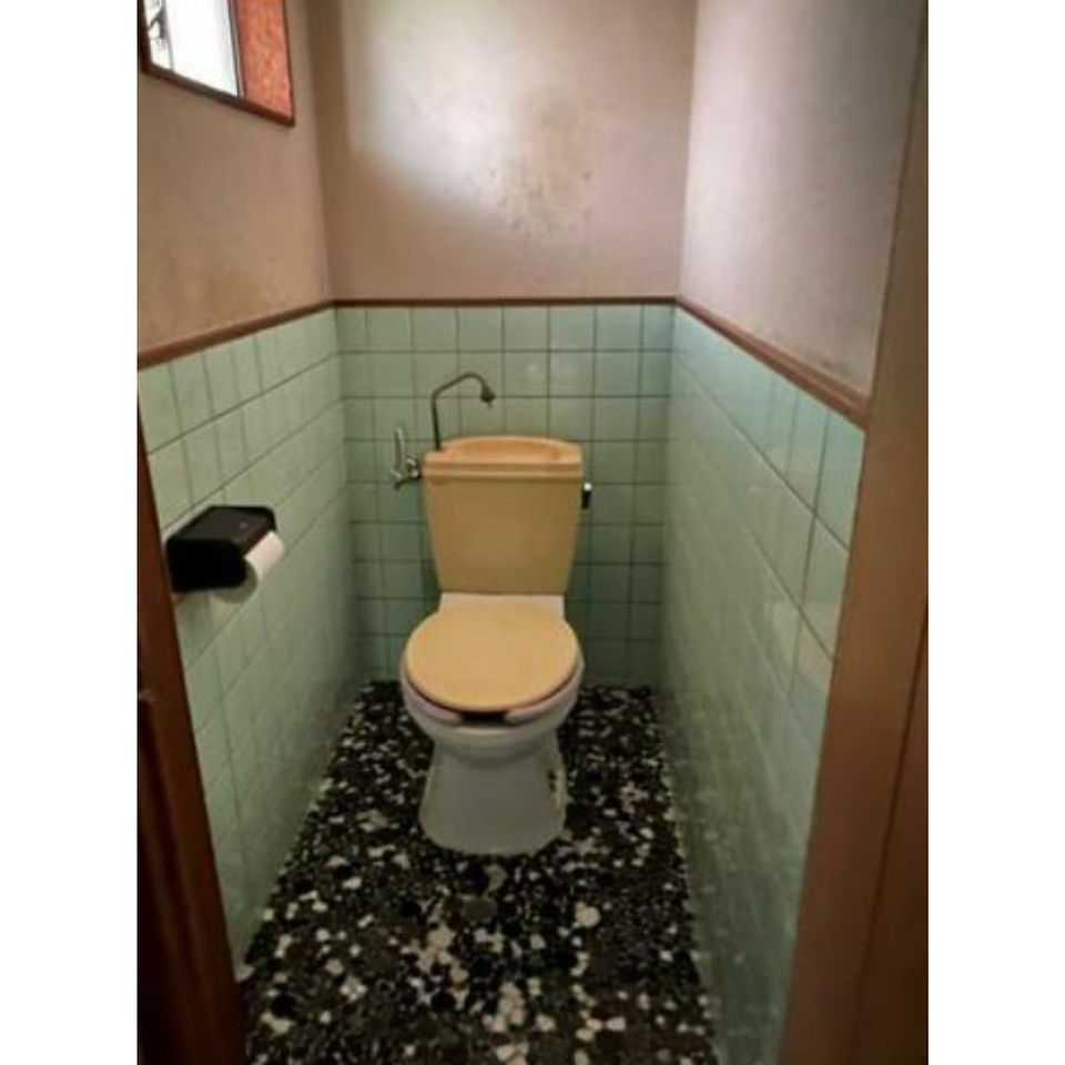 The photograph depicts a small, old-fashioned toilet room with green tiles on the walls. The toilet itself is a vintage beige color with a matching lid. A small sink is attached to the wall above the toilet, and a toilet paper holder is mounted on the adjacent wall. The floor is covered in small, black and white tiles.