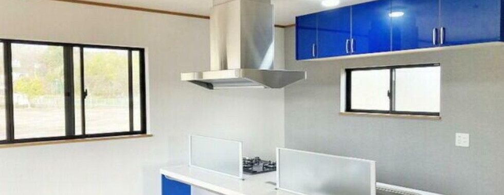 A photo of a modern kitchen with blue cabinets and a white countertop. The kitchen features a stainless steel range hood, a gas stove, and a double sink. There is a glass partition separating the cooking area from the dining area. The kitchen has large windows with blinds and the floor is a light-colored wood.