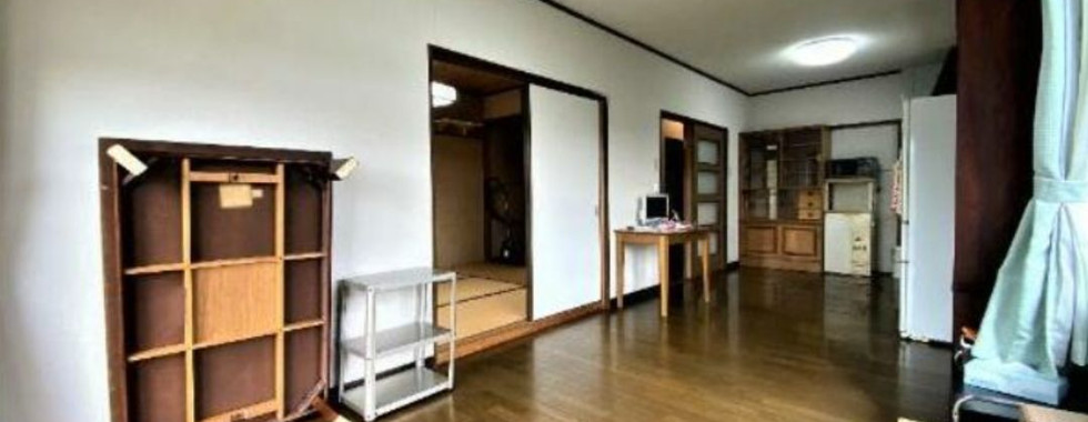 The photograph shows a spacious living and dining room area with wooden flooring and white walls. A traditional Japanese-style dining table (chabudai) is placed in the center of the room. In the background, there is a glimpse of a kitchen and a hallway leading to other rooms.