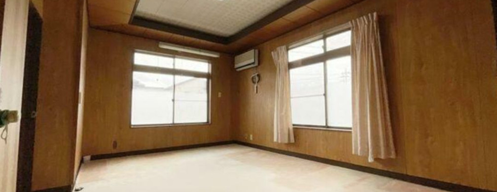 The image shows a large, empty room with wooden walls and a carpeted floor. There are two windows on one wall, providing natural light. The room has a simple and functional design, with no furniture or decorations.