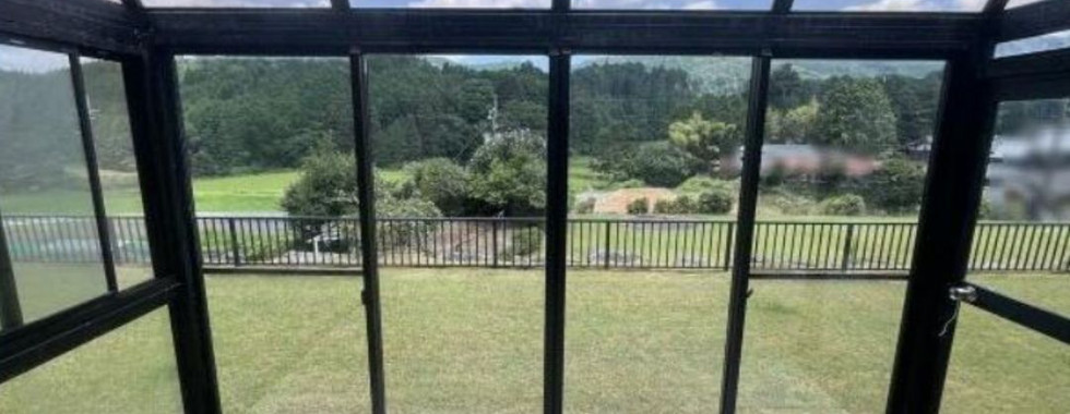 The photo shows a sunroom with a glass roof and walls. The room offers a panoramic view of a green landscape with rolling hills and trees. The floor is made of wooden planks, and there are several windows and doors providing access to the outdoors. The overall impression is of a bright and airy space perfect for enjoying the natural surroundings.