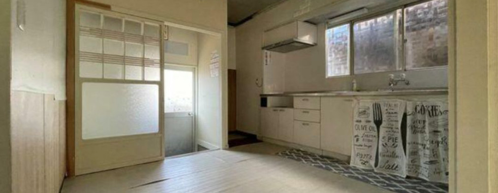 An open-plan kitchen and dining room area in a Japanese house. The kitchen features light-colored cabinets and a countertop with a sink. A window above the sink provides natural light. The dining area has wood flooring and shoji sliding doors that lead to another room.