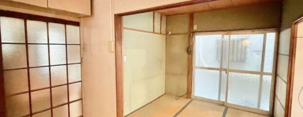 The photo shows a Japanese-style room with tatami mats covering the floor. There is a sliding door with a frosted glass panel leading to another room on the left side. On the right side, there is a sliding glass door with a clear glass panel leading to a balcony or outdoor area. The walls are covered in a light-colored wood paneling.