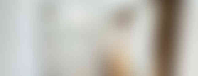 A photo of a front entranceway with a white door and a wooden shoe rack with a mirror above it. The walls are painted white and the floor is tiled. There is a window with blinds in the background.