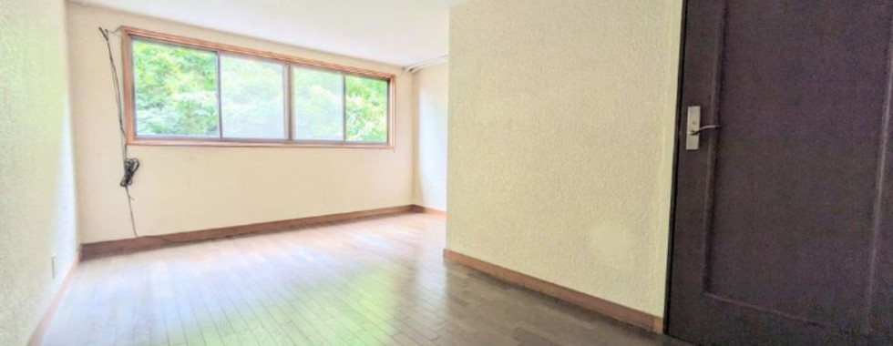 Empty bedroom with wooden floors and a large window overlooking greenery. A door is visible on the right wall.