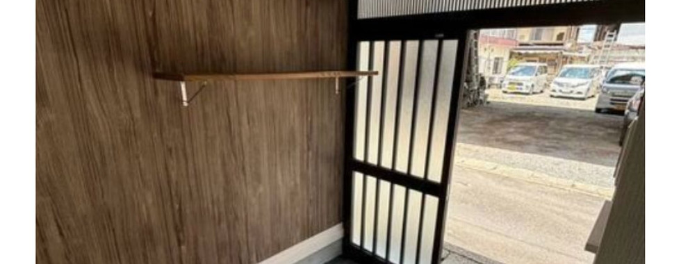 This photo shows the entranceway to a home. There is a wooden shelf mounted on a wall with a wood-like texture. Below the shelf, there is a small step leading up to the main floor. The entrance door is open, revealing a view of a street with parked cars.