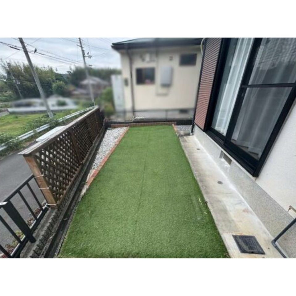 This photo shows a small backyard with artificial grass. The yard is bordered by a wooden fence and a brick wall. There is a small concrete patio area along the house, and a black gate on the left side of the image.