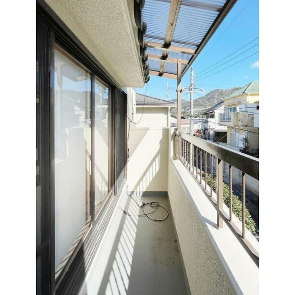 The photo shows a balcony on a sunny day. The balcony has a concrete floor and is covered by a wooden pergola. There is a metal railing and a window to the left. The view from the balcony includes a residential area with houses and a mountain in the distance.