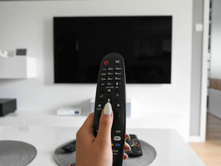 A hand holds a remote control, pointing at a blank black TV screen. The room is modern with a white and gray color scheme.