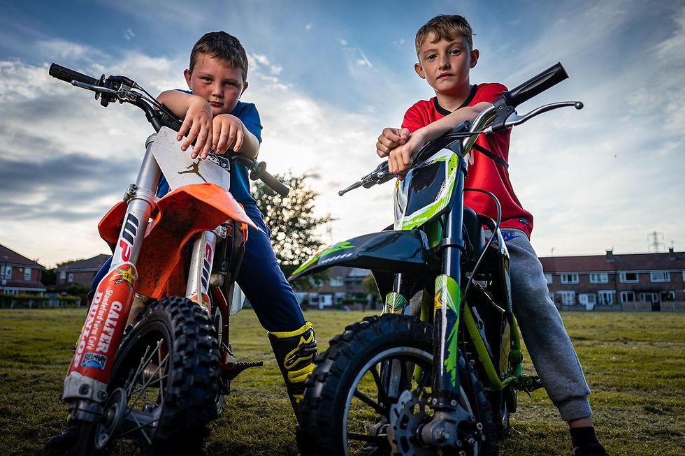 Two boys pose on dirt bikes in a grassy field at sunset, with houses and a cloudy sky behind them.