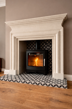 Black Heta Inspire 45 wood-burning stove with visible flame, installed in a light stone fireplace surround with patterned tile interior and decorative tiled hearth.