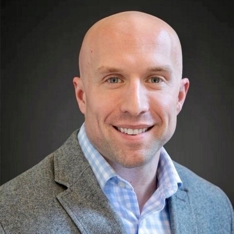 Smiling bald man wearing a grey blazer and blue checkered shirt.