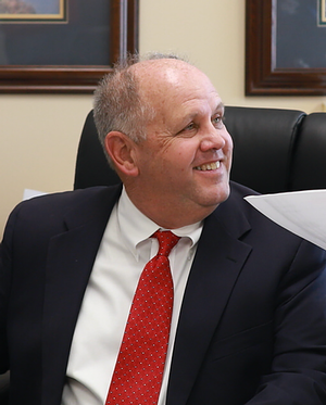 Mark R. Conway “Jr.” smiling in a suit and red tie.