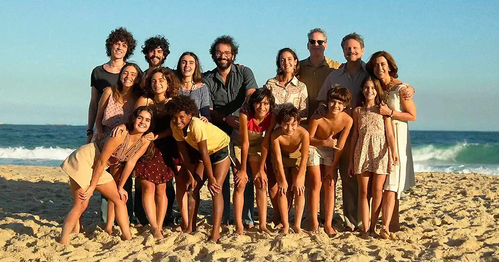 Um grupo de 15 pessoas, incluindo adultos e crianças, posa sorridente para uma foto em uma praia, com o mar e o céu azul ao fundo. As pessoas estão vestindo roupas leves e coloridas, algumas de vestido, outras de shorts e camisetas. A cena transmite alegria e cumplicidade, com um clima ensolarado e descontraído.