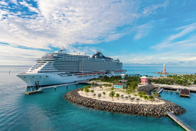 Navio de cruzeiro atracado em uma ilha tropical com águas azul-turquesa, céu parcialmente nublado, vegetação com palmeiras, pequenas construções coloridas e um farol listrado ao fundo.