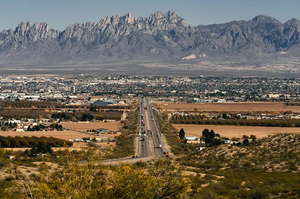 Paisagem urbana com uma estrada reta cortando a imagem em direção a uma cidade ao fundo, cercada por áreas agrícolas e vegetação desértica. Ao fundo, destaca-se uma imponente cadeia de montanhas rochosas sob céu claro, provavelmente localizada no sudoeste dos Estados Unidos.