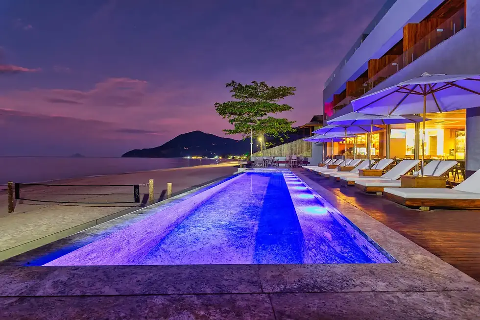 Alt text: Piscina iluminada em tons de azul à beira-mar ao entardecer, com um hotel de luxo ao lado, espreguiçadeiras sob guarda-sóis e vista para montanhas ao fundo. O céu exibe tons de roxo e laranja enquanto as luzes do hotel criam um ambiente acolhedor.