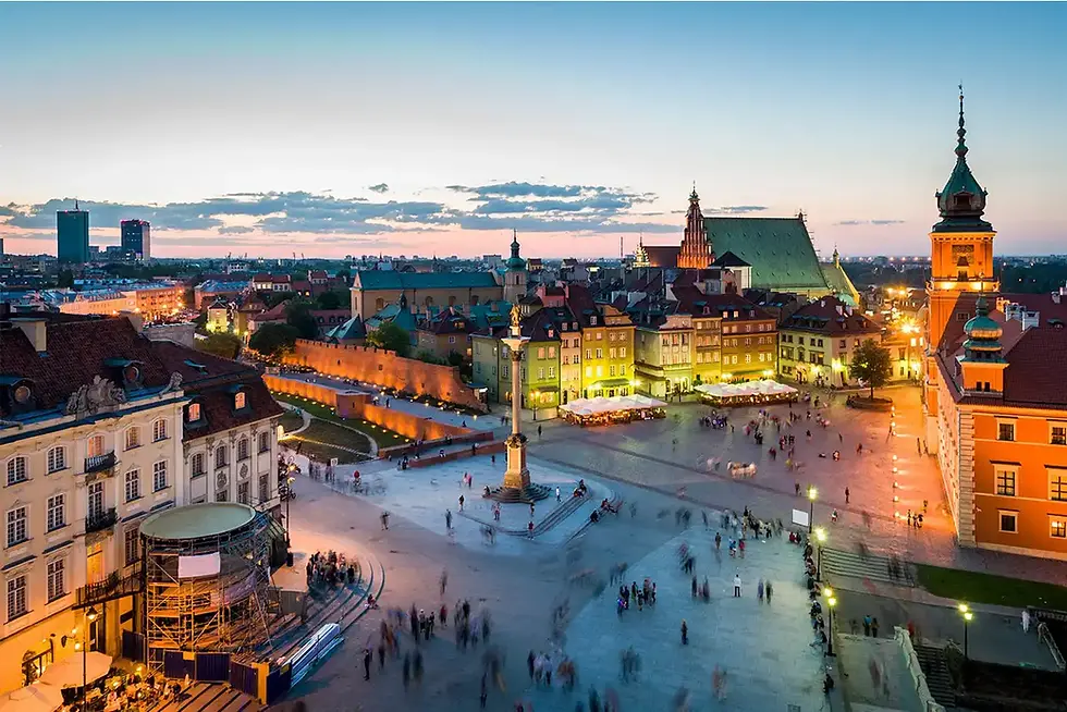 Vista panorâmica da Praça do Castelo em Varsóvia, Polônia, ao entardecer. A imagem mostra a praça movimentada com pessoas caminhando, a Coluna de Sigismundo no centro e edifícios históricos iluminados ao redor. No fundo, destaca-se o Castelo Real de Varsóvia com sua torre característica e a Catedral de São João. O céu exibe tons alaranjados e azuis, criando uma atmosfera charmosa na cidade.