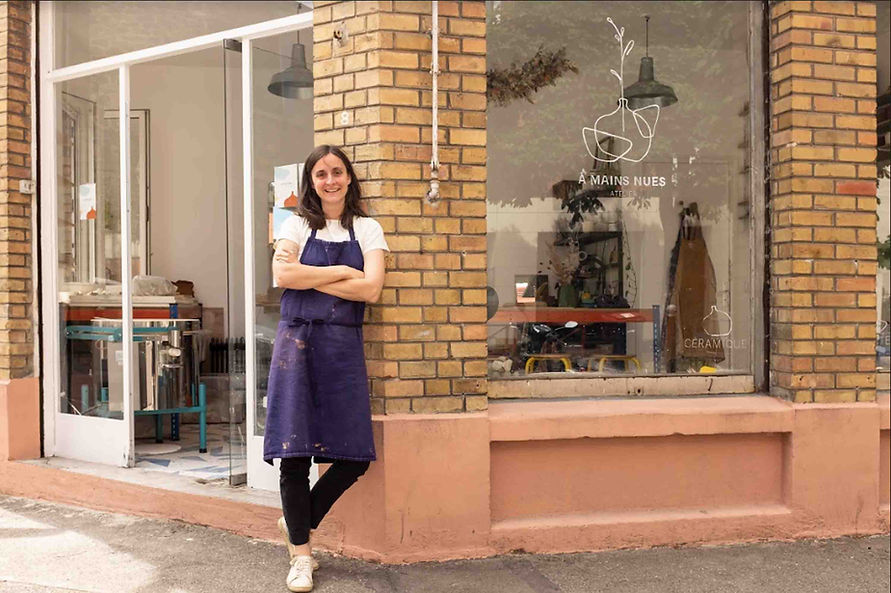 Une femme céramiste se tient devant son atelier de poterie. Elle porte un tablier, a les bras croisés et sourit.