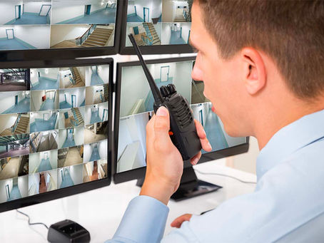 A security personnel monitoring multiple CCTV feeds on screens while using a walkie-talkie, showcasing advanced remote monitoring capabilities.