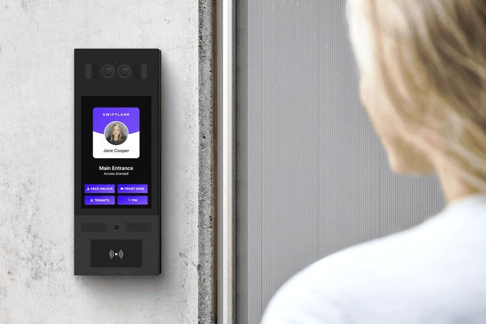 A close-up view of a sleek, black digital intercom and access control panel (labeled 'Swiftlane') mounted on a wall, showing a visitor interface. A person is seen from behind approaching the device. This represents modern Virtual Doorman technology.