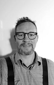 Man wearing glasses smiles, suspenders visible, against white background. Portrait shot.