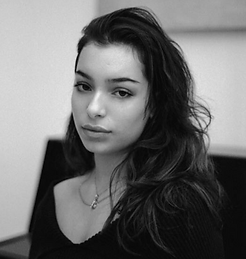 Young woman with dark wavy hair and a pendant stares directly at camera.