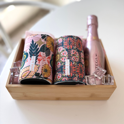 A small bamboo tray styled in soft pink tones with two patterned Bask & Sip drink sleeves and a mini bottle of sparkling; tied with a simple ribbon.
