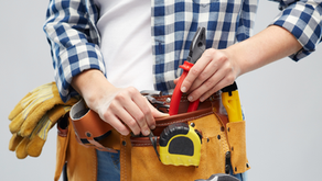 Person in a blue checkered shirt wearing a tool belt with pliers, gloves, and a tape measure, standing against a plain background.