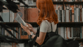 Red-haired woman in a white shirt reads a book in a library. Bookshelves in the background, dark foliage partially obscuring view. Calm atmosphere.