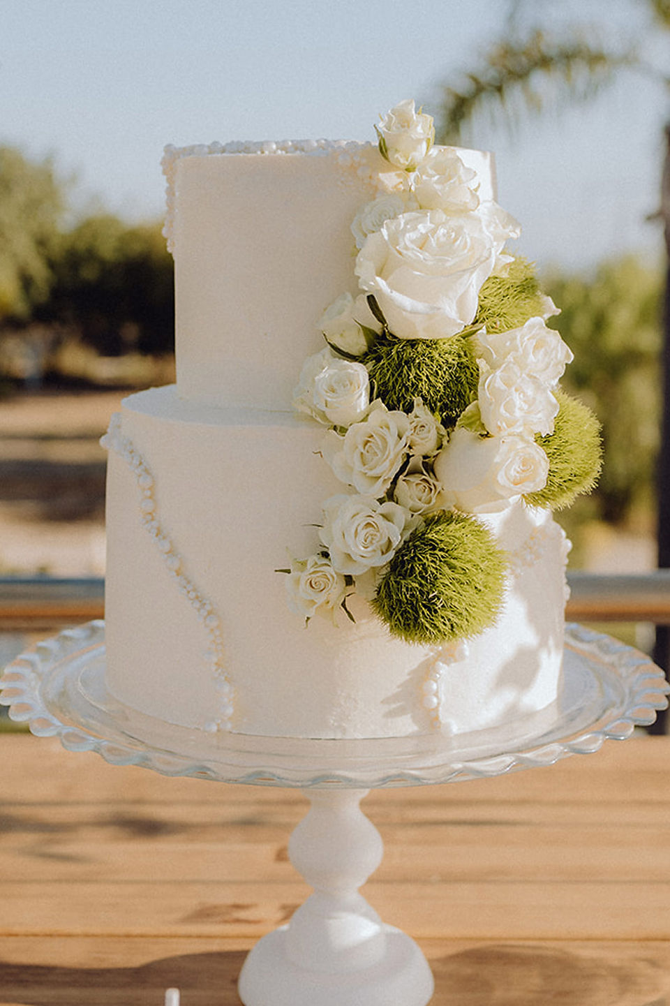 Bolo de casamento branco de dois andares com flores brancas e verdes em arranjo floral lateral.