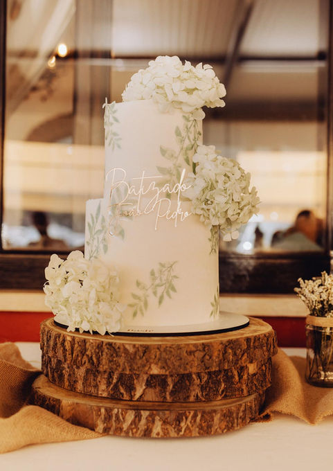 Bolo de batizado elegante em branco com flores brancas, folhagem verde e decoração delicada