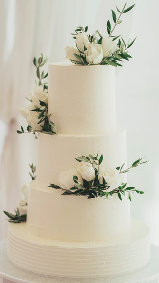 Bolo de casamento branco de três andares com flores brancas e ramos verdes em decoração elegante.