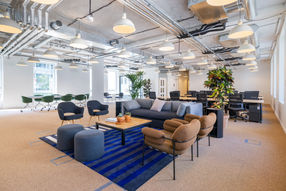 Vue d’un espace de coworking moderne avec des sièges confortables, des luminaires suspendus et des éléments de design industriel, avec des tables de réunion en arrière-plan et des plantes d’intérieur.