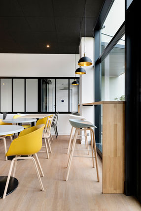 Café moderne avec chaises jaunes, tabourets hauts, et grandes fenêtres.