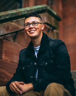 A young man sits on the footsteps of a brick building smiling