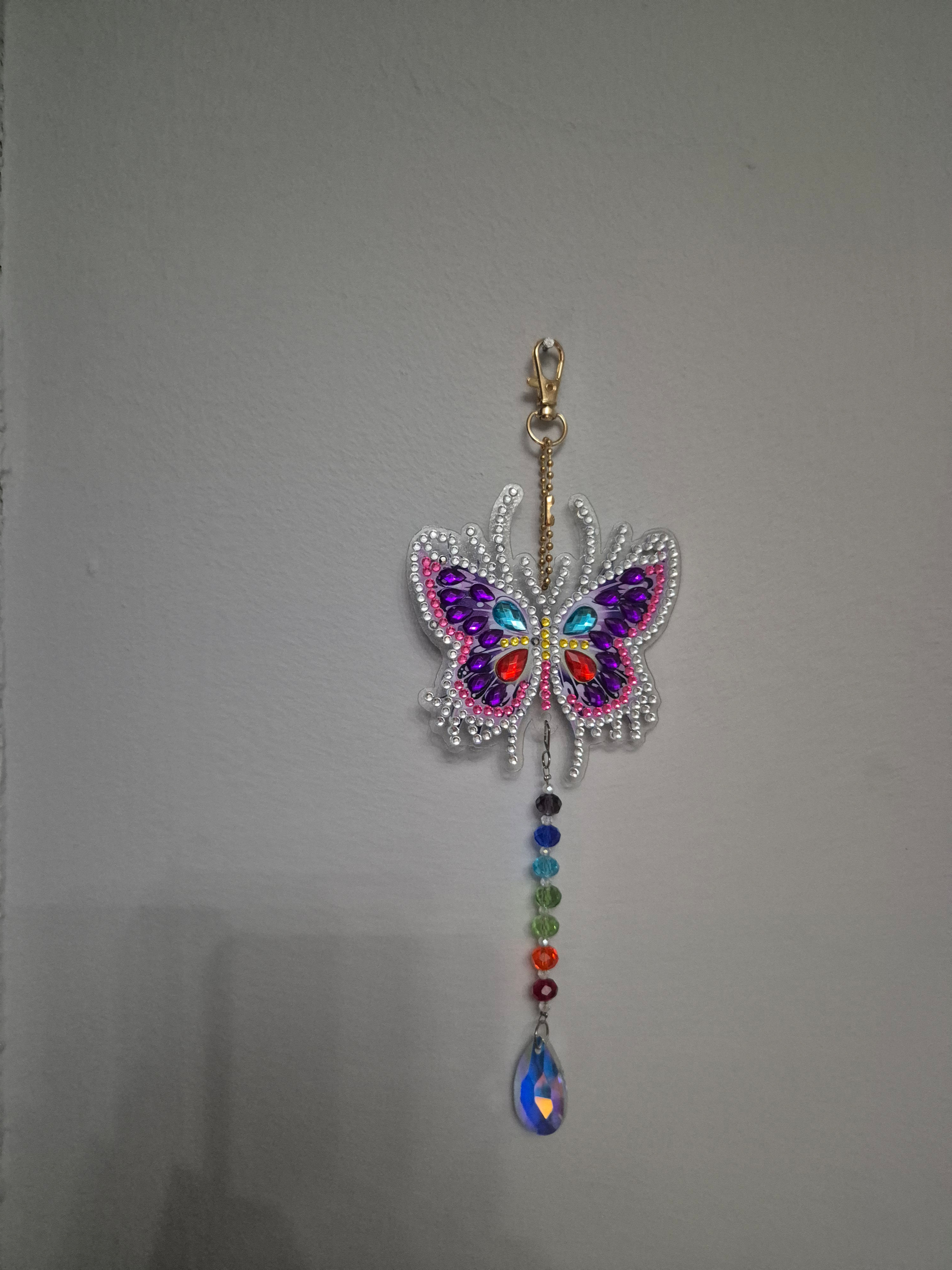 A bejeweled butterfly ornament with colorful crystals, hanging against a gray wall.