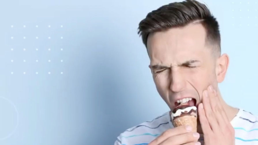 Man in striped shirt biting ice cream cone, wincing in pain while touching cheek. Light blue background with dotted pattern.