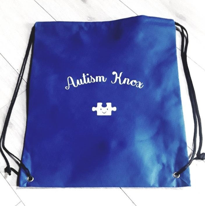 Blue drawstring bag that says "Autism Knox" with a white puzzle piece that has a smiley face on it below the writing. Available for purchase through website. 