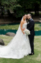 Bride in a white gown and groom in a black suit embrace, holding white flowers by a pond in a green, leafy park setting.