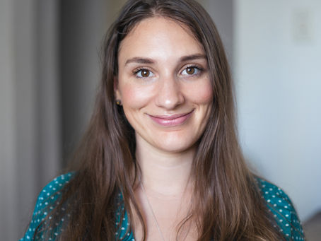 A woman with long brown hair and a teal polka-dot top smiles at the camera. She is wearing a silver necklace and is positioned in a softly lit indoor setting.