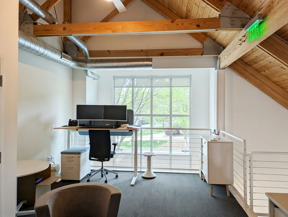 Standing desk workstation with dual monitors in the HELD Architecture remodel for VS America in Charlotte, NC, featuring a black ergonomic chair, exposed wood beam ceilings, large windows with natural light and green views, and polished concrete floors