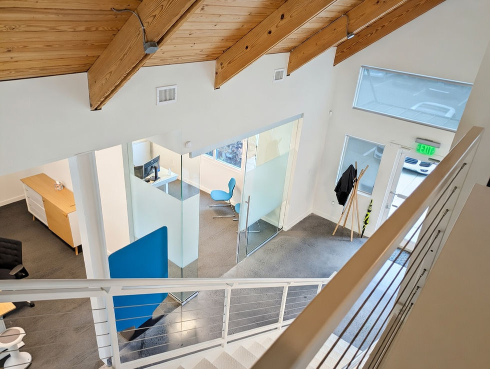 Staircase overlook of the modern open office interior designed by HELD Architecture for VS America in Charlotte, NC, featuring glass partitions, a bright blue chair, exposed wood beam ceilings, natural light, and polished concrete floors.