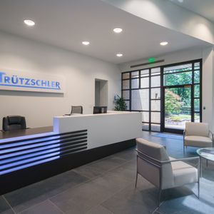 Reception area at American Trützschler with branded front desk, soft seating, circular glass coffee tables, and views to landscaped exterior through tall windows.