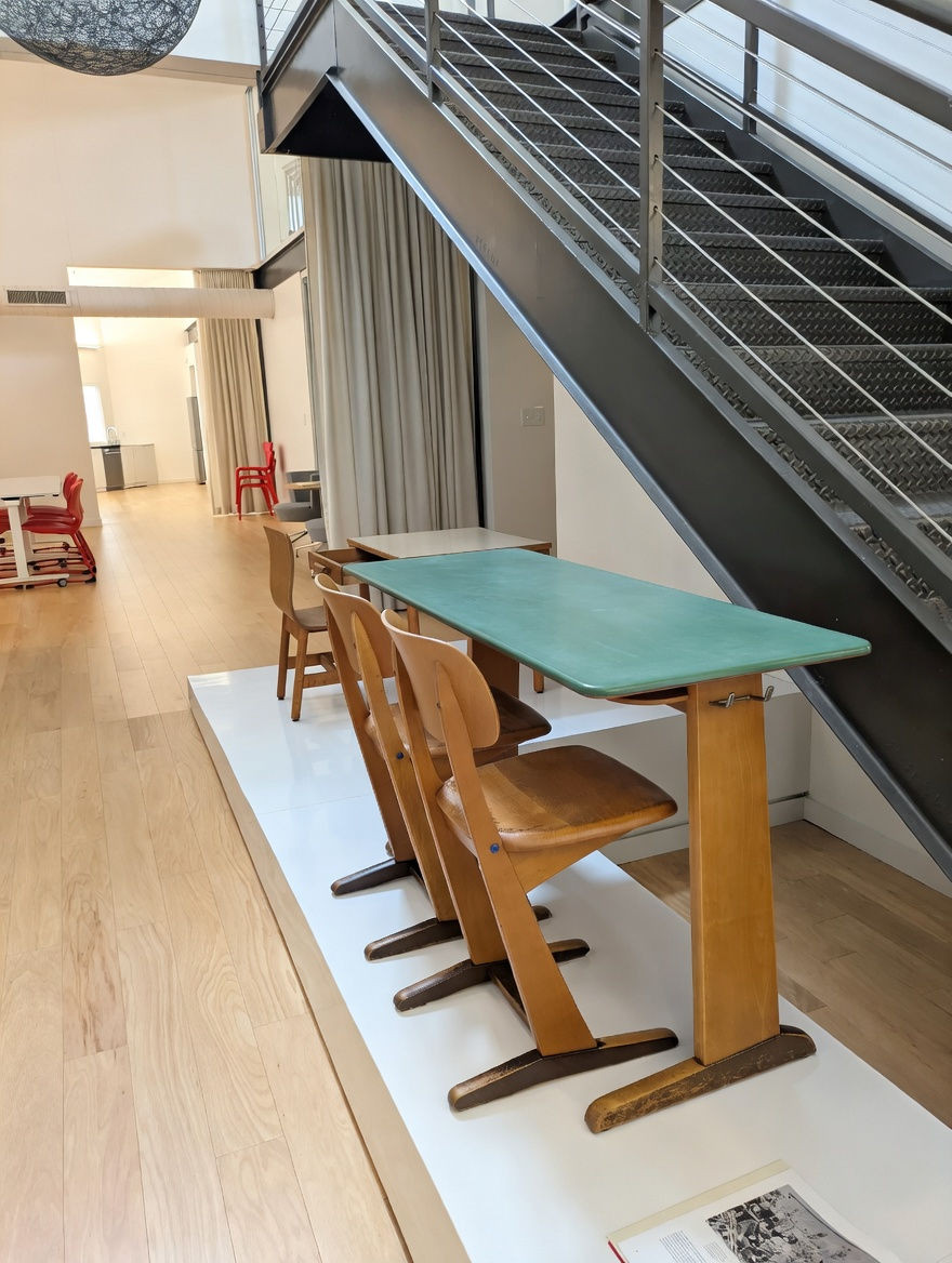 Wooden school chairs and green table display designed by HELD Architecture for VS America office and showroom renovation in Charlotte, NC, featuring classic ergonomic furniture on a white platform beneath a modern staircase with exposed wood ceilings.